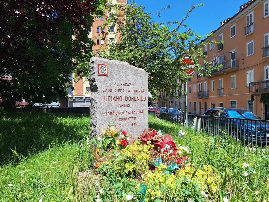 Cippo commemorativo dedicato a Luciano Domenico, decorato con fiori e circondato da edifici e vegetazione.