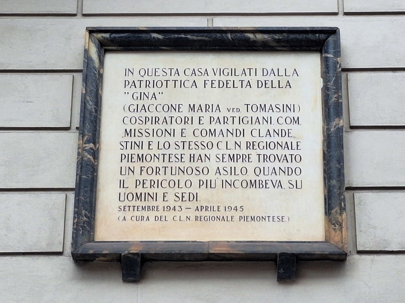 Lapide in marmo con cornice scura, fissata su parete esterna, con lunga iscrizione commemorativa.