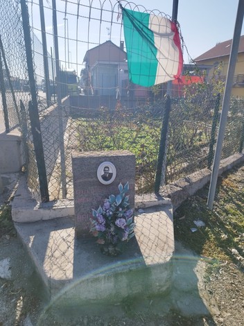 Cippo commemorativo dedicato a Barisone, con fiori e bandiera italiana sullo sfondo in un'area recintata.