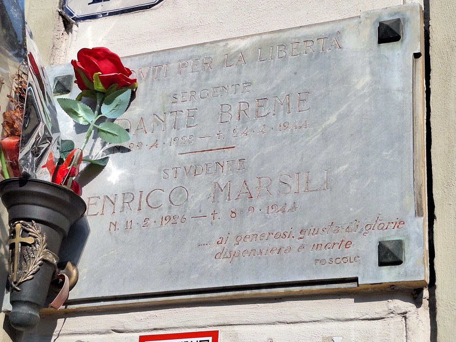 Lapide in marmo su parete esterna, con iscrizione e rosa rossa deposta davanti.