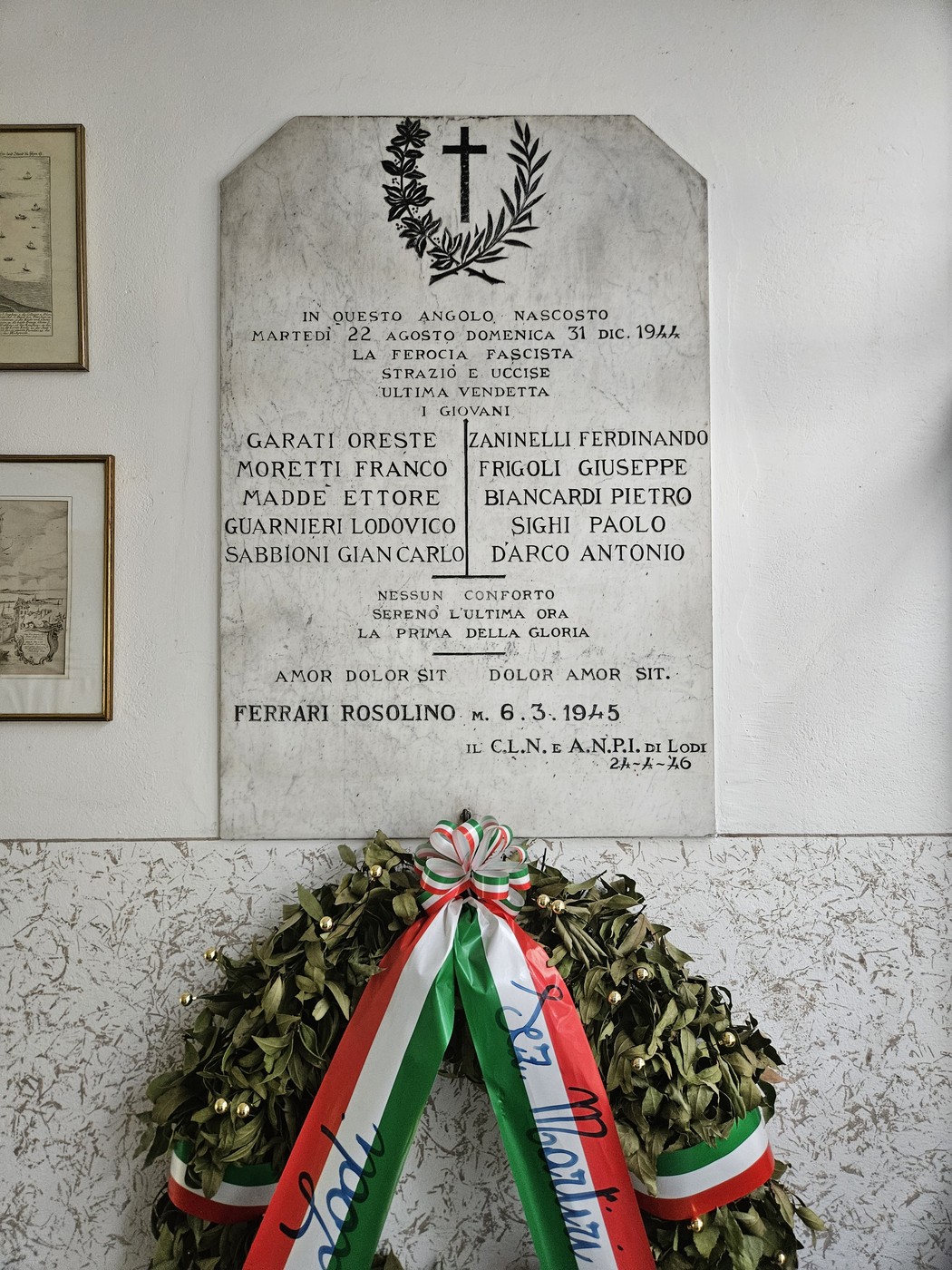 Lapide in marmo con croce scolpita e iscrizione, appesa a parete, sotto una corona floreale con nastro tricolore.