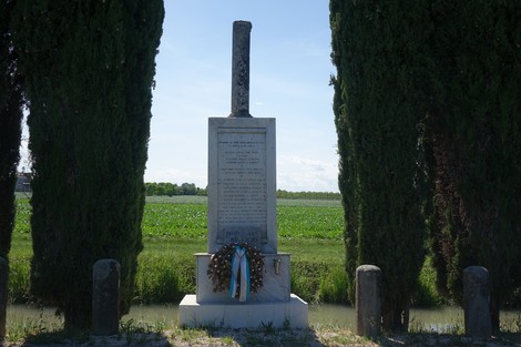 Monumento commemorativo dedicato a Gennaro con una corona di fiori, circondato da alberi e campo verde sullo sfondo.