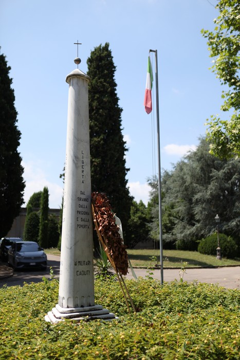 Monumento ai caduti con obelisco bianco, corona di alloro e bandiera italiana sullo sfondo.
