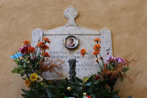 Lapide commemorativa di Pierino Caminada, decorata con fiori e un'immagine centrale, su fondo murario.