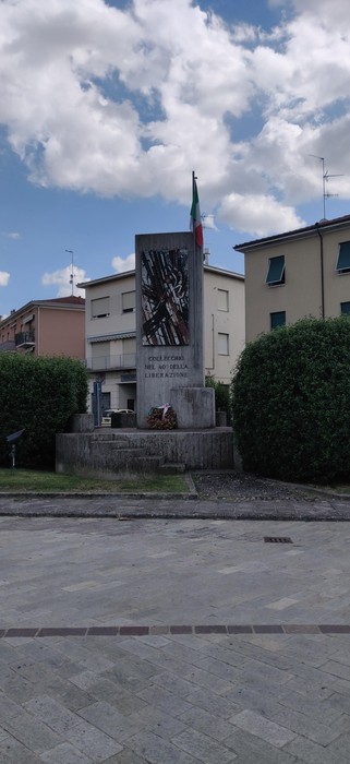 Monumento ai partigiani con bandiera italiana, situato in un'area cittadina recintata da siepi verdi.