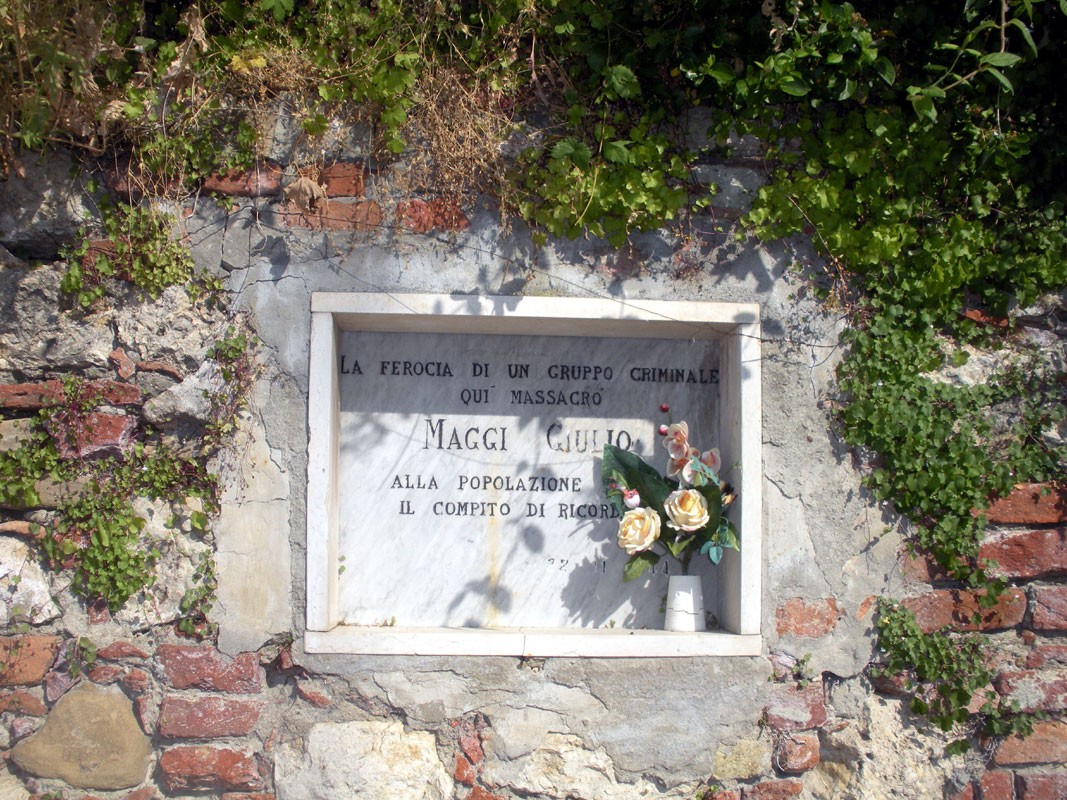 Lapide commemorativa dedicata alle vittime di un massacro avvenuto a Maggi, circondata da fiori e vegetazione.