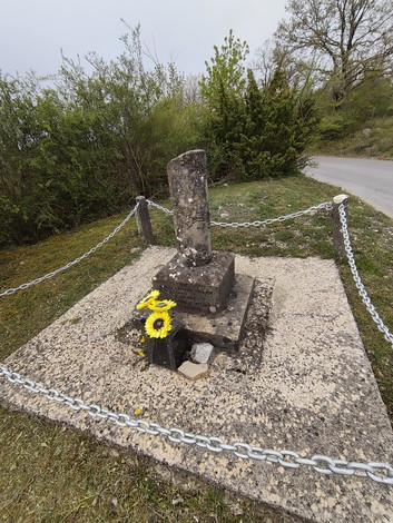 Cippo commemorativo a Ulivelli circondato da catene e fiori, immerso nella natura.