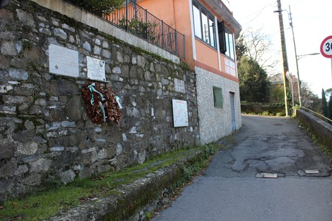 Lapide commemorativa su un muro di pietra, circondata da foglie e in un contesto rurale. La strada sale verso l'orizzonte.