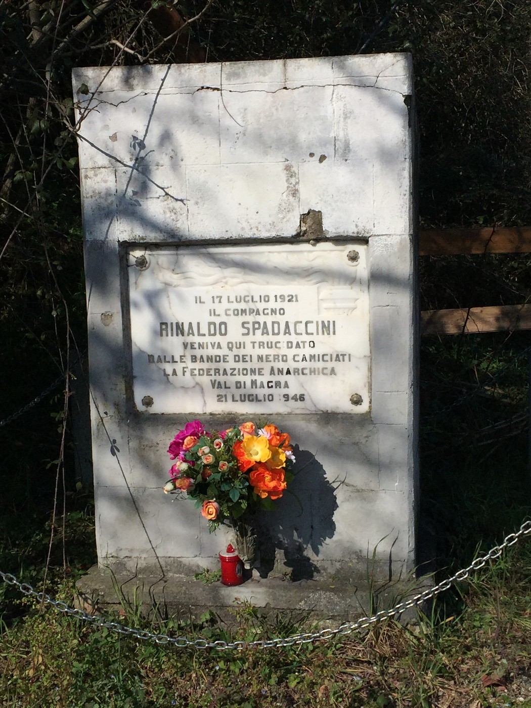 Cippo commemorativo dedicato a Rinaldo Spadaccini, con fiori e candele, situato nella natura.