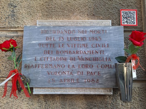 Lapide in pietra con iscrizione su parete, ai lati rose rosse e un portafiori metallico.