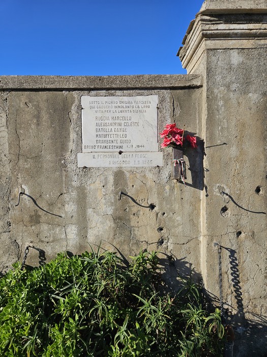 Lapide commemorativa dedicata ai Martiri della FLAGE, con decorazioni floreali e parete di pietra.