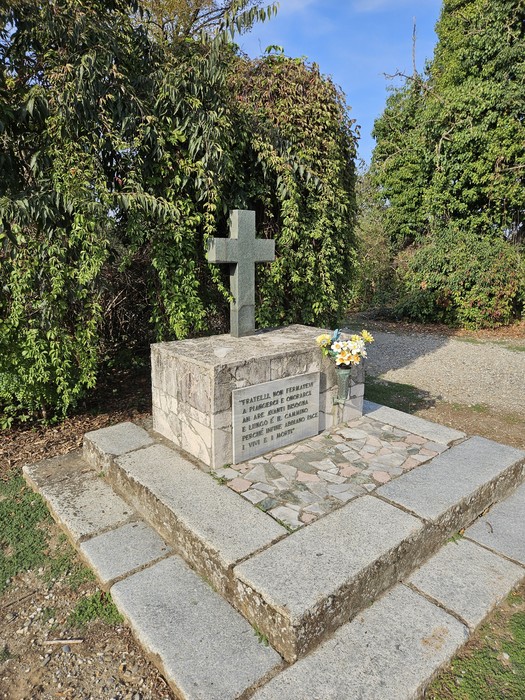 Monumento in pietra con croce, base con iscrizione e fiori, su basamento tra alberi e siepi.