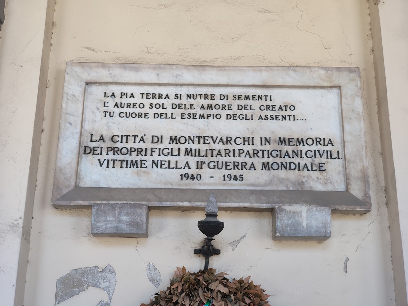 Lapide marmorea con iscrizione, fissata su una parete, con mensola e corona di foglie sottostante.