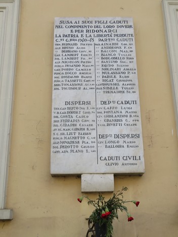 Lapide commemorativa dedicata ai caduti per la patria, con nomi e date incisi su pietra. Fiori ai piedi della lapide.