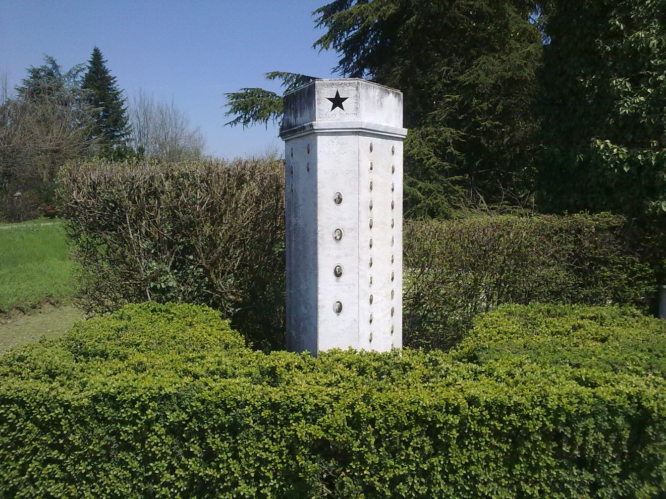 Monumento ai Caduti di San Biagio, circondato da siepi verdi e alberi, in un contesto naturale.