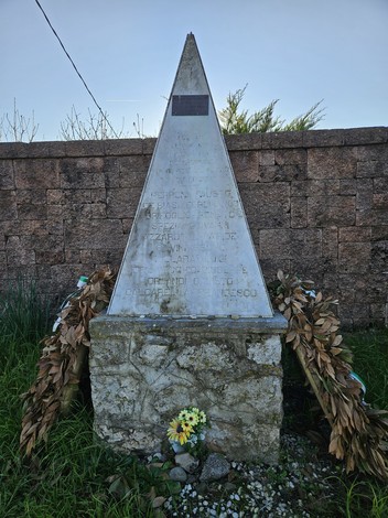 Monumento ai Martiri di Ressora, obelisco con decorazioni floreali e un muro di pietra sullo sfondo.