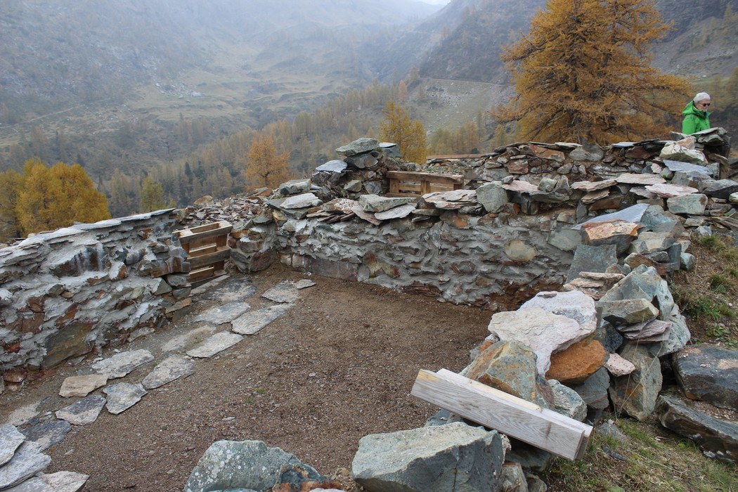 Ruderi del "Vecchio rifugio dei Laghi Gemelli" circondati da natura e montagne, esposizione di pietre e legno.