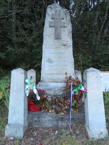 Monumento a Graziotti con croce in pietra, fiori e decorazioni, immerso nella vegetazione.