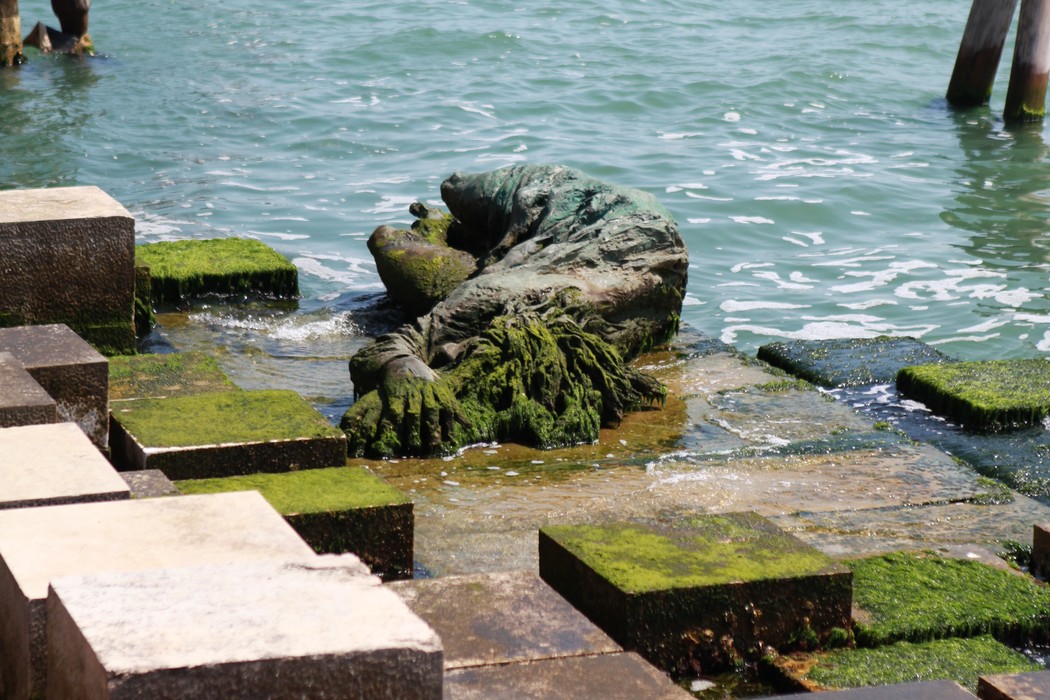 Monumento alla partigiana immerso nella natura, con vegetazione che cresce attorno alla statua e acqua sullo sfondo.