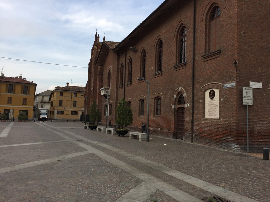 Lapide a Olivelli, monumento commemorativo in una piazza storica, circondato da edifici e alberi.