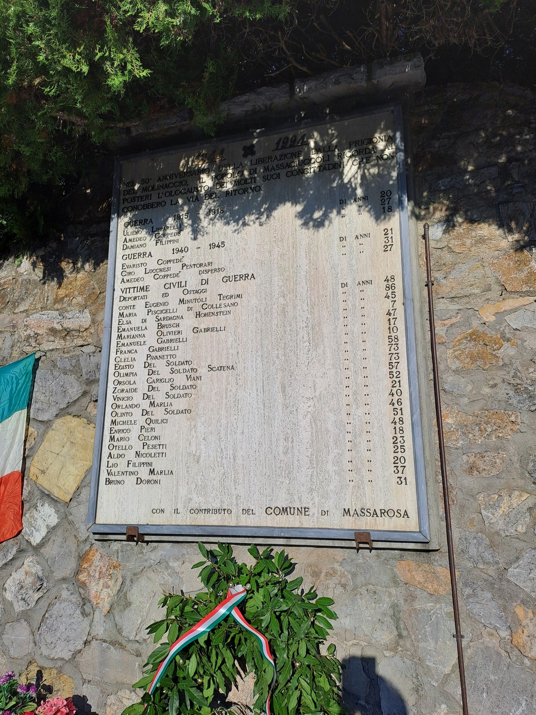 Lapide in marmo con iscrizione, fissata a un muro in pietra e decorata con una corona di alloro.