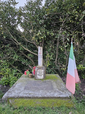Cippo commemorativo con decorazioni floreali e bandiera italiana, circondato da una vegetazione verde.