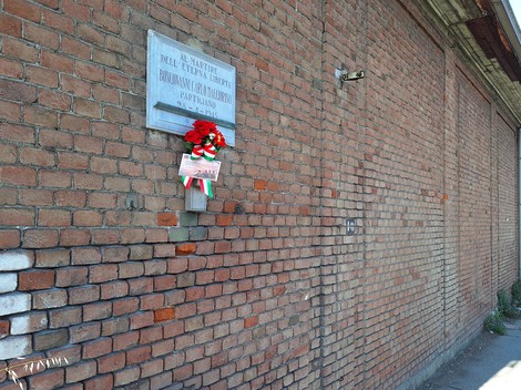 Lapide commemorativa su muro di mattoni, adornata con una corona di fiori e un nastro tricolore.