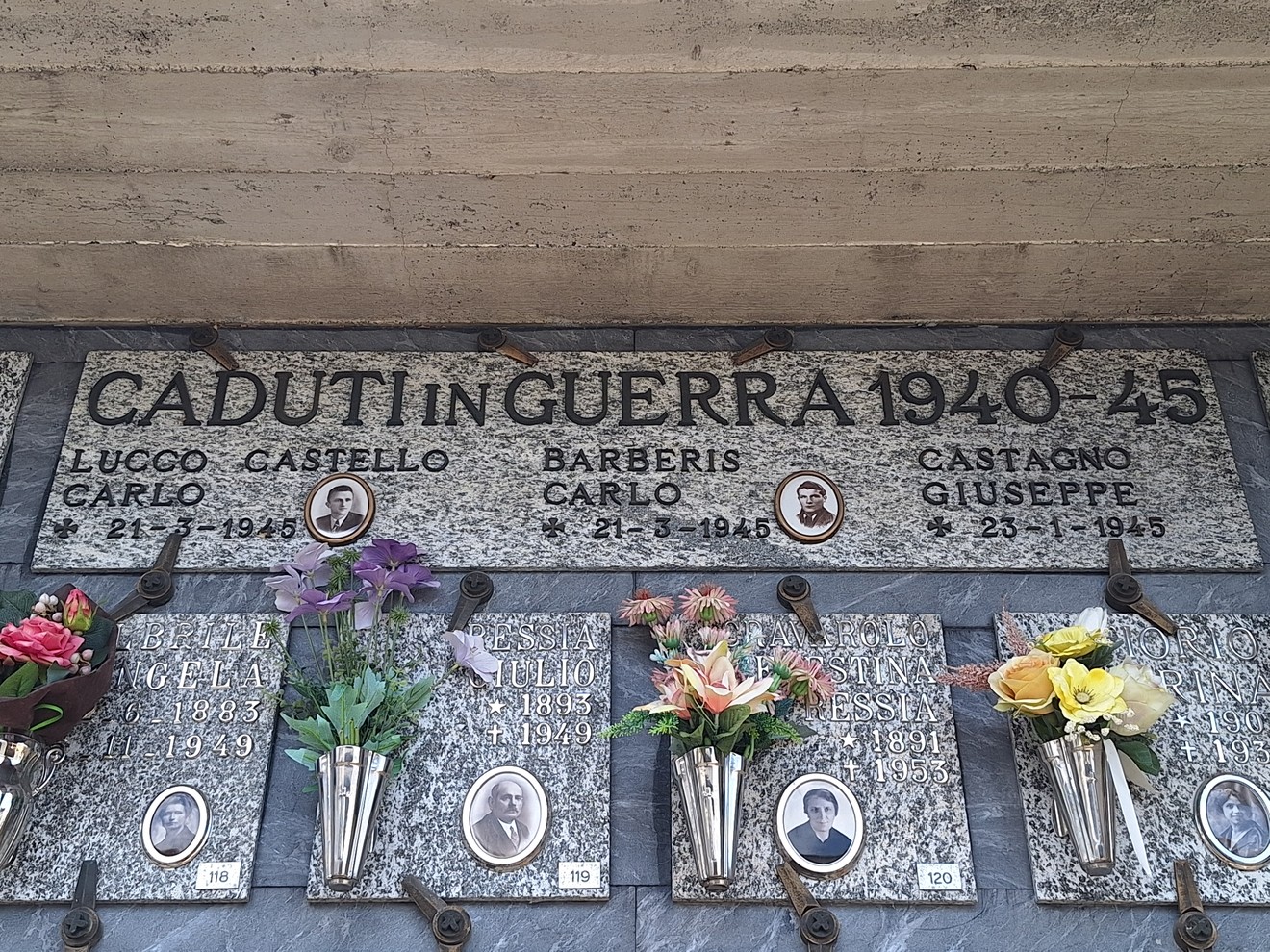 Lapide in marmo con iscrizione e fototessere dei caduti, sotto fiori in vasetti metallici alla parete.