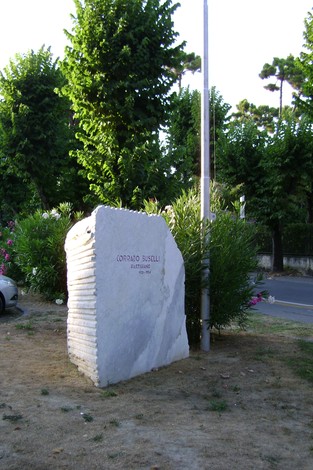 Cippo commemorativo dedicato a Corrado Buselli, situato in un'area verde con alberi e piante.