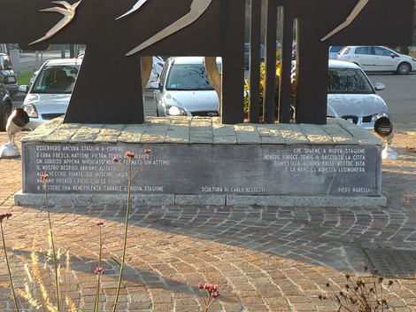 Monumento in pietra con iscrizione, sormontato da sagome metalliche di rondini, su una piazza con auto parcheggiate.