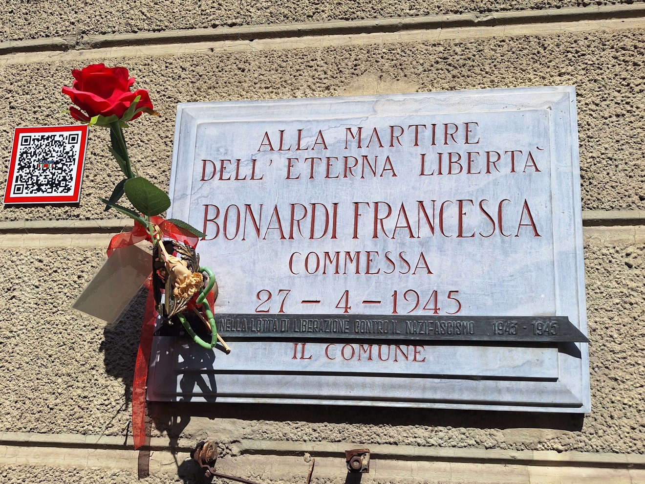 Lapide in marmo su parete con iscrizione, una rosa rossa e un mazzo di fiori appesi sotto.