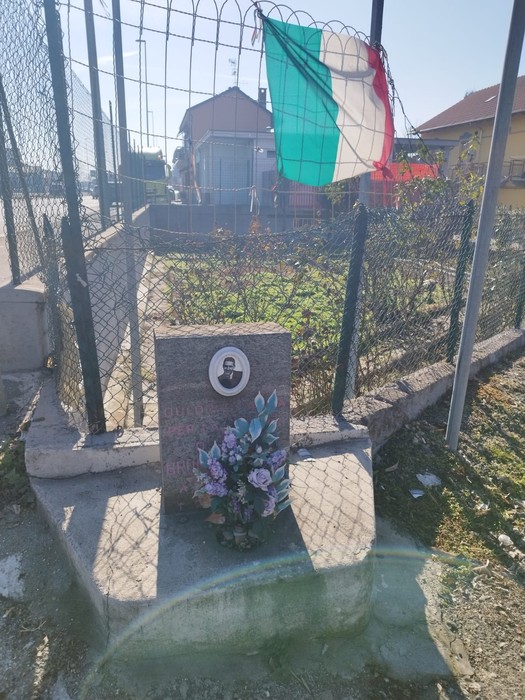 Cippo commemorativo dedicato a Barisone, con fiori e bandiera italiana sullo sfondo in un'area recintata.
