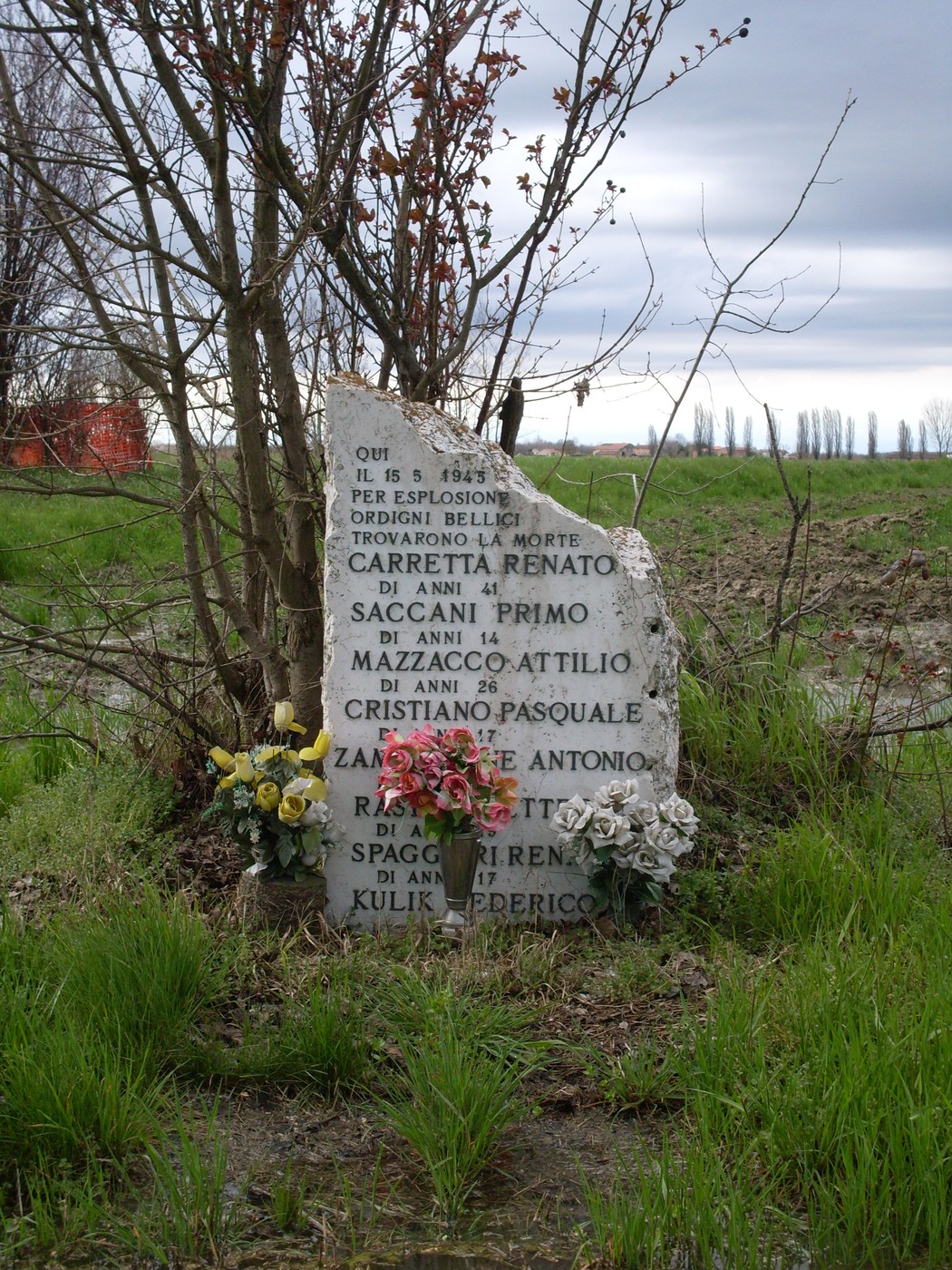 Cippo commemorativo dedicato alle vittime di un bombardamento, decorato con fiori e circondato da vegetazione.