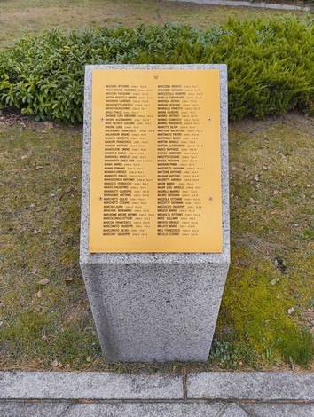 Targa commemorativa con nomi di caduti nel Cimitero militare italiano di Zehlendorf, circondata da vegetazione.