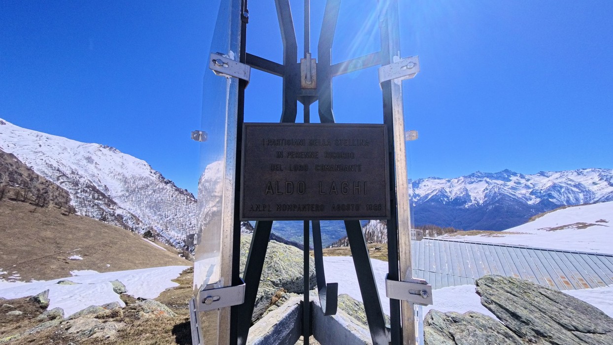 Targa commemorativa con iscrizione su struttura metallica in quota, circondata da montagne innevate.