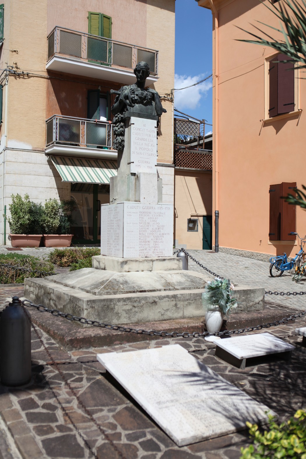 Monumento ai Caduti con statua e lastre commemorative in un'area urbana, circondato da edifici e vegetazione.