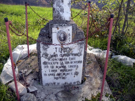 Cippo commemorativo con iscrizioni e ritratto, circondato da erba verde e catene. Ambiente rurale.