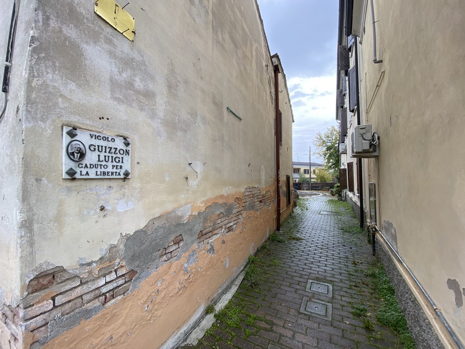 Vicolo stretto con una lapide commemorativa dedicata a Guizzon, circondato da edifici storici.
