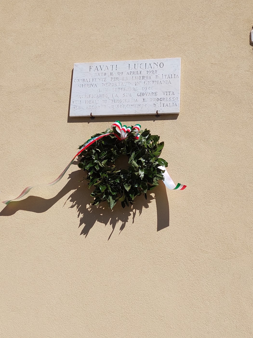 Lapide commemorativa a Luciano Favati, con una corona di alloro e nastri tricolori, affissa su una parete.