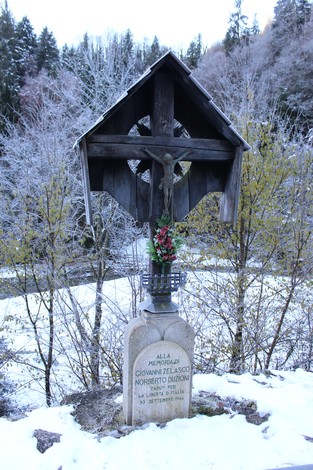 Croce monumentale dedicata a Giovanni Zelasco, situata in un paesaggio innevato e boscoso.