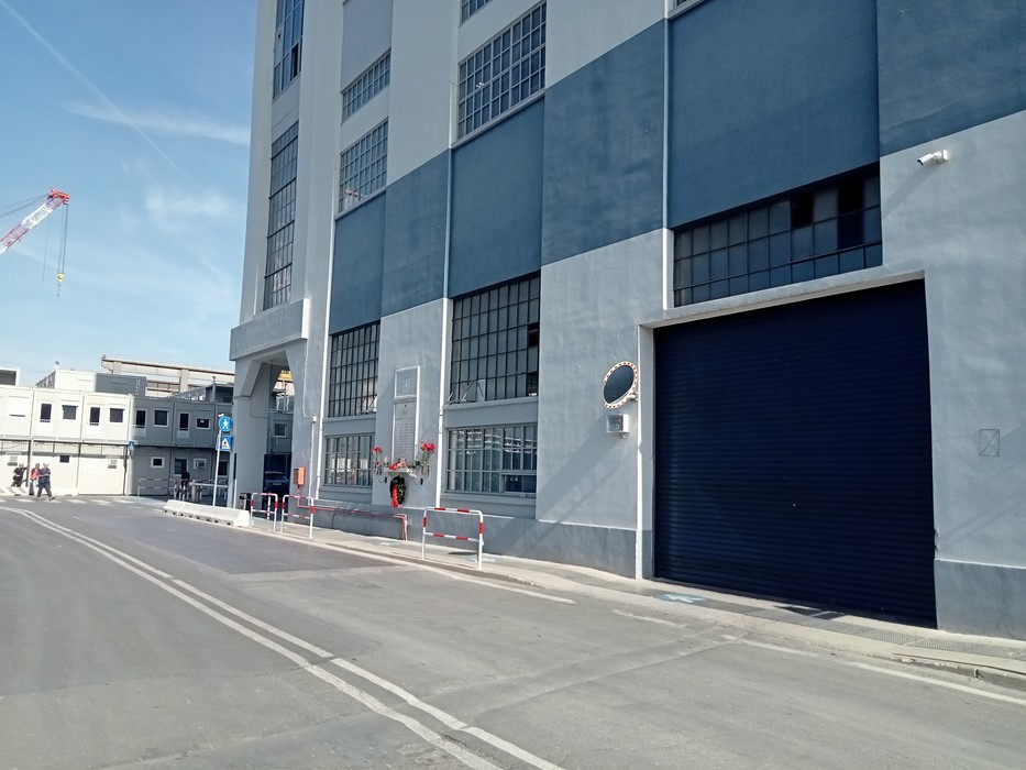 Facciata di uno stabilimento industriale con targa commemorativa accanto a un ingresso con saracinesca.