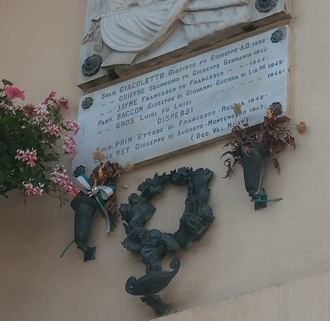 Lapide commemorativa con iscrizioni, decorata da fiori e una corona, in memoria dei caduti.
