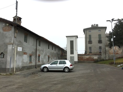 Lapide commemorativa dedicata alla Piazza XXV Aprile, con edifici storici sullo sfondo e un'auto parcheggiata.