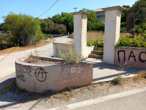 Lapide commemorativa in un'area rurale, circondata da vegetazione e segni di degrado urbano.
