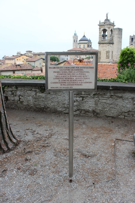 Targa commemorativa dedicata a Quasimodo, con vista su un paesaggio urbano e edifici storici.