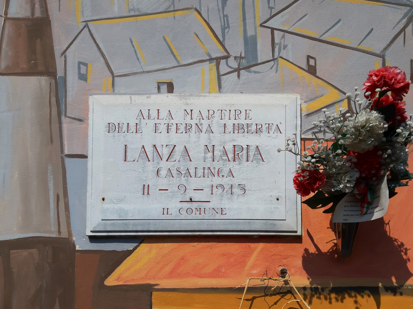 Lapide commemorativa dedicata a Lanza Maria, con fiori, situata in un'area decorata da murales.
