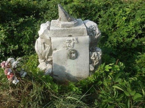 Cippo commemorativo immerso nella vegetazione, con decorazioni floreali e un elemento appuntito sulla sommità.