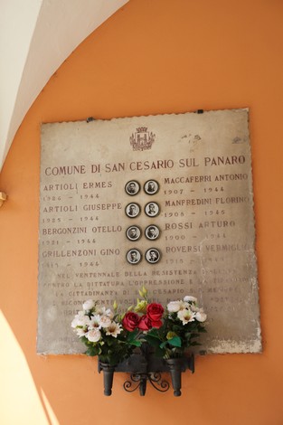 Lapide commemorativa ai partigiani caduti, con nomi e date incisi su pietra, adornata da fiori.