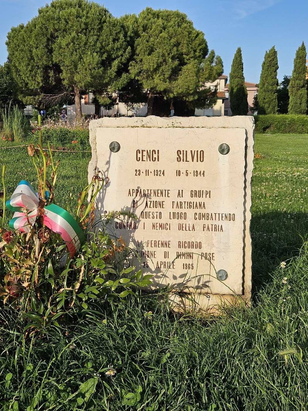 Cippo commemorativo dedicato a Silvio Cenci, situato in un'area verde, circondato da piante e decorazioni tricolori.