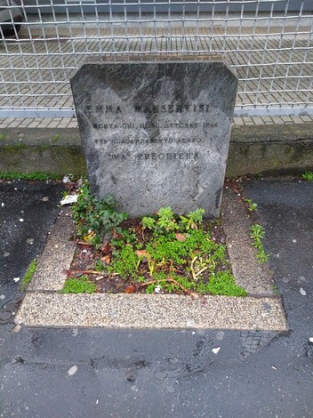 Cippo commemorativo dedicato a Emma Manservisi, con flora circostante e ambiente urbano.